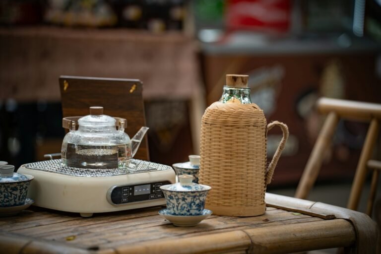 A glass teapot on an electric stove beside a wicker-wrapped bottle and blue and white tea cups