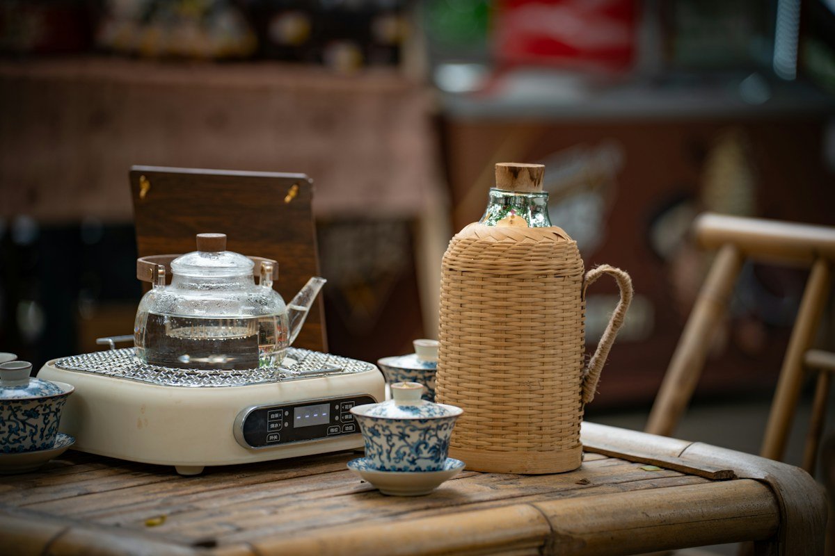 A glass teapot on an electric stove beside a wicker-wrapped bottle and blue and white tea cups